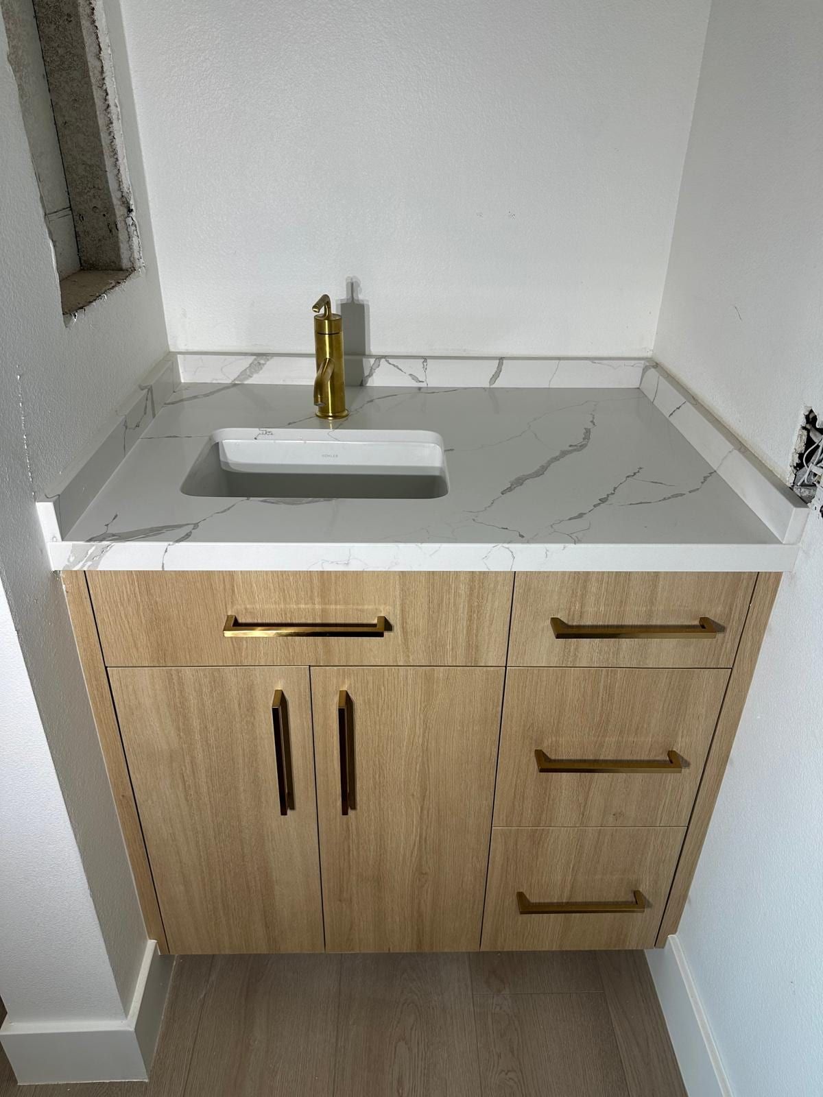 A light oak vanity with brass pulls, a white quartz top, an undermount sink and a brass faucet