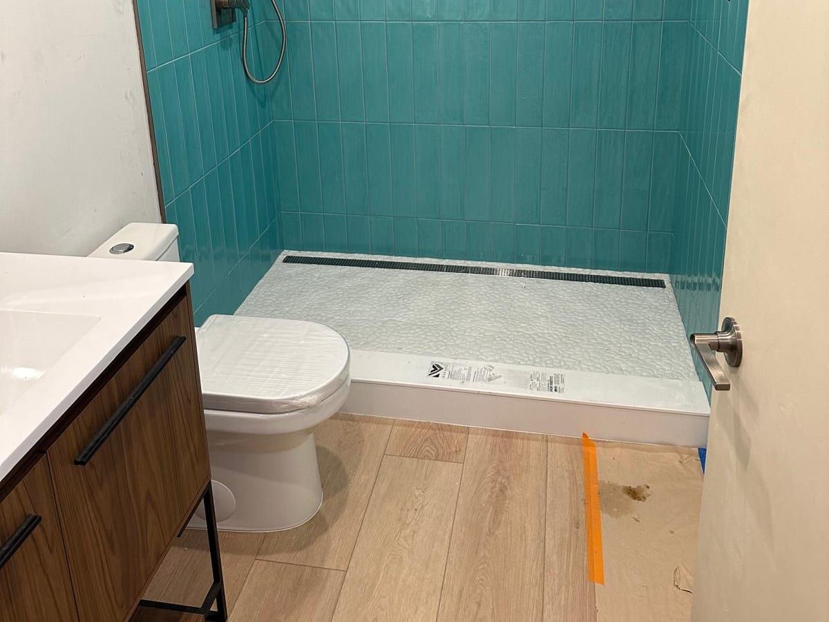A new toilet beside a walnut vanity, facing an aqua tiled walk-in shower with a linear drain